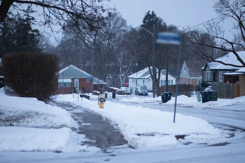 Residential street after a snowfall, with snow piled along the sidewalks and bungalows beyond.
